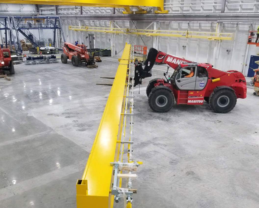 http://Workers%20using%20a%20Manitou%20telehandler%20to%20position%20a%20large%20yellow%20overhead%20crane%20beam%20inside%20an%20industrial%20facility