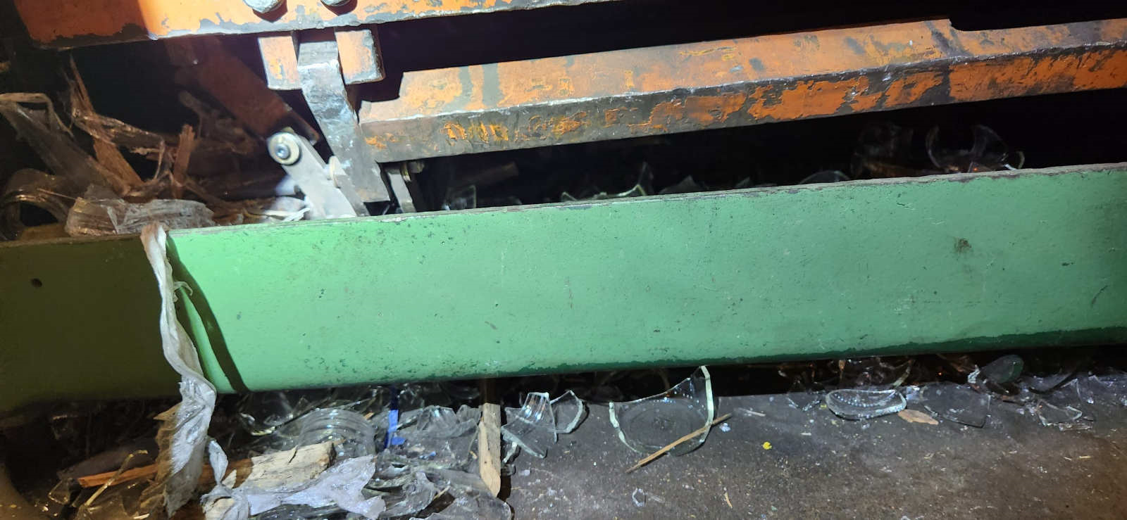 Close-up of a conveyor or press system with broken glass and debris in a maintenance area.